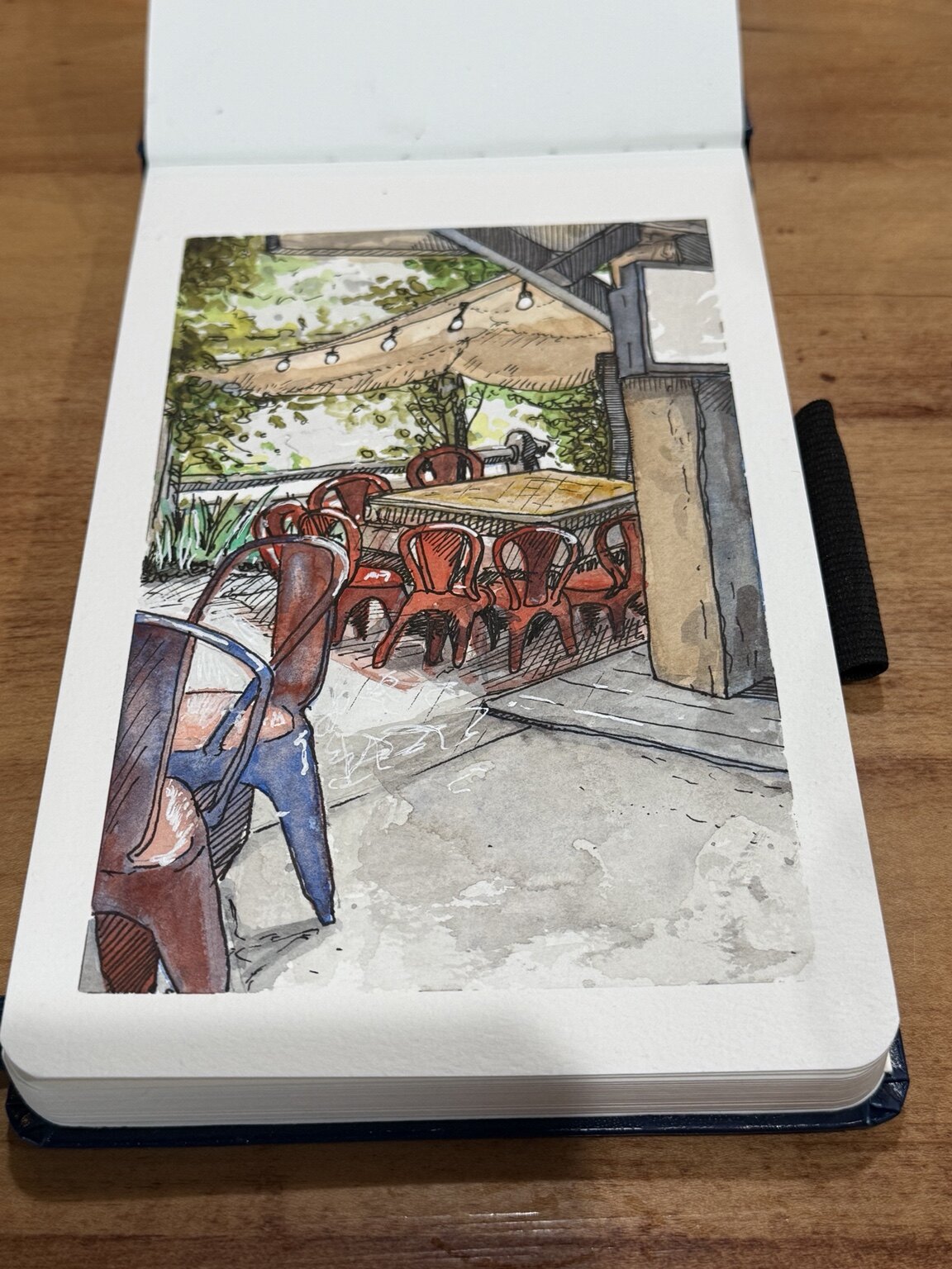 Watercolor and ink sketchbook painting of red and blue chairs gathered around tables on a shaded patio.